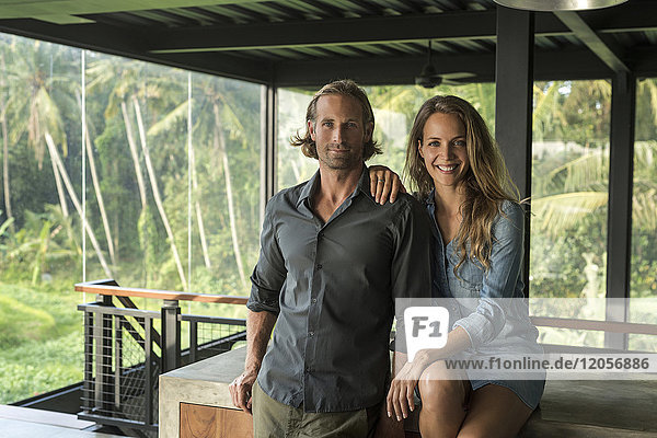 Paar lächelnd vor der Kamera in einem zeitgenössischen Designhaus mit Glasfassade  umgeben von einem üppigen tropischen Garten.