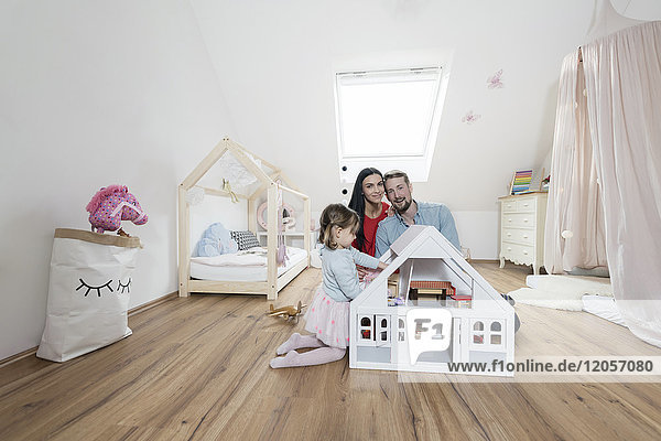 Glückliche Eltern im Kinderzimmer  während die kleine Tochter mit einem Puppenhaus spielt.
