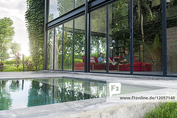 Gartenblick durch Glasfassade des Paares mit Laptop und Buch auf roter Couch in modernem Designhaus vor dem Schwimmbad