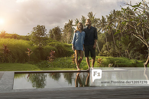 Lächelndes Paar steht am Rande eines Pools in einem üppigen tropischen Garten