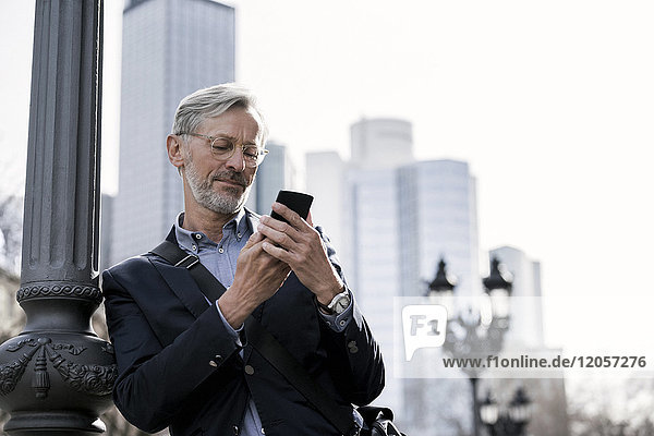 Grauhaariger Geschäftsmann mit Blick auf das Smartphone neben der Straßenlaterne