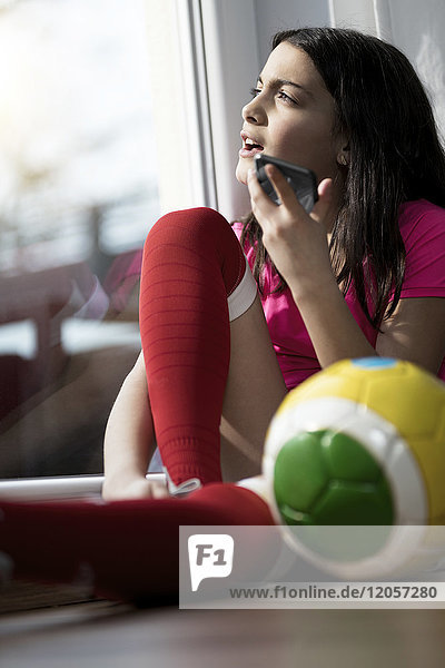 Mädchen im Fußball-Outfit sitzt auf dem Boden im Wohnzimmer und spricht mit jemandem auf ihrem Smartphone.