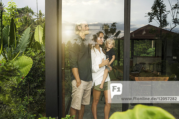 Gartenansicht der Eltern mit ihrem kleinen Sohn  der außerhalb ihres Designhauses inmitten eines üppigen tropischen Gartens blickt.