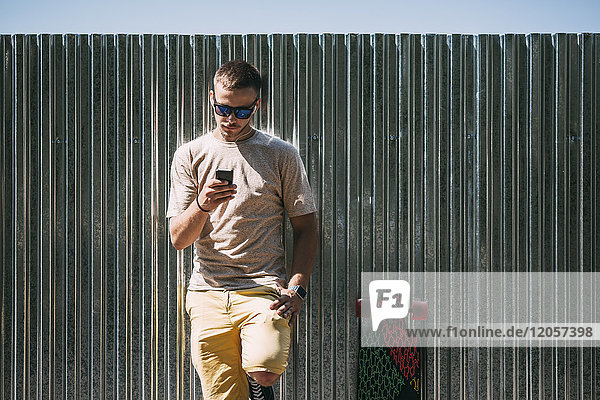 Junger Mann mit Handy und Ohrstöpseln  die an einer Wand neben dem Longboard lehnen