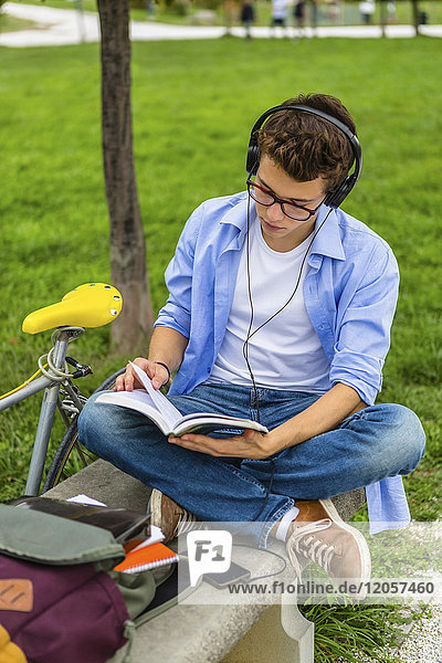 Young man with racing cycle and headphones sitting on a bench reading book