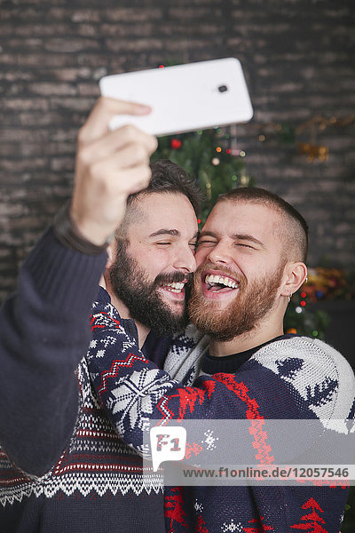 Porträt eines lachenden schwulen Paares  das sich zu Hause mit dem Smartphone in die Weihnachtszeit begibt.
