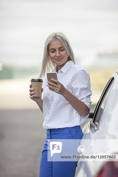 Smiling businesswoman with takeaway coffee and cell phone outside car
