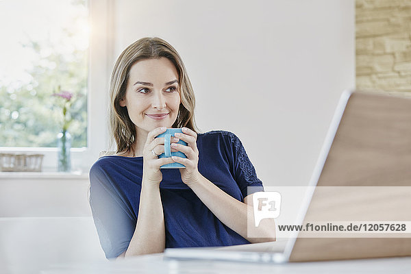 Smiling woman at home with coffee mug and laptop