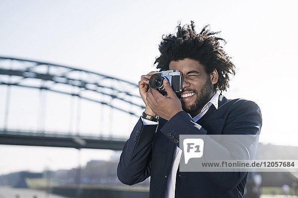 Lächelnder Mann im Anzug am Flussufer beim Fotografieren mit einer Vintage-Kamera