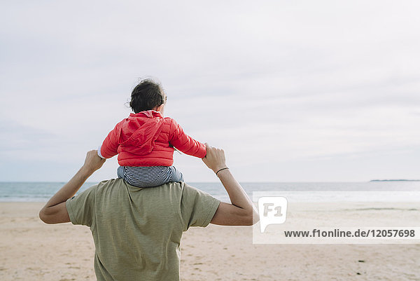 Rückansicht des Vaters  der seine kleine Tochter auf den Schultern am Strand trägt.
