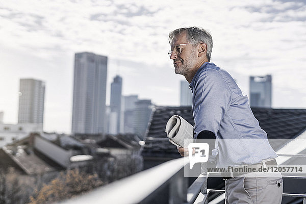 Grauhaariger Mann steht auf dem Balkon mit Zeitungsausschnitt über der Stadt