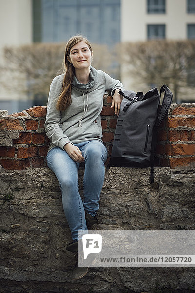 Porträt einer lächelnden jungen Frau  die mit ihrem Rucksack an der Wand sitzt.