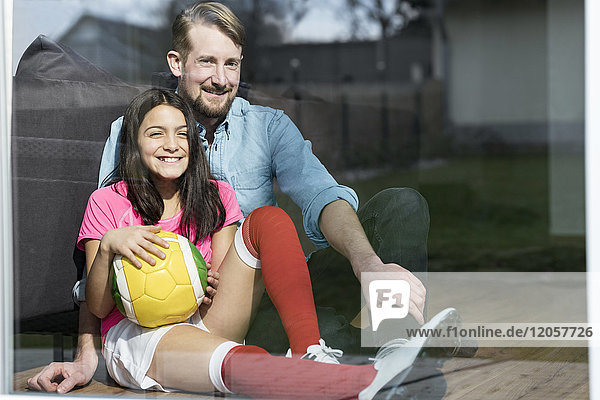 Girl in soccer outfit sitting next to father on floor in living room looking out window