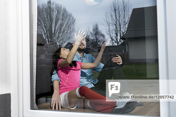 Mädchen im Fußball-Outfit sitzt neben Vater auf dem Boden im Wohnzimmer und spielt mit dem Fußball.
