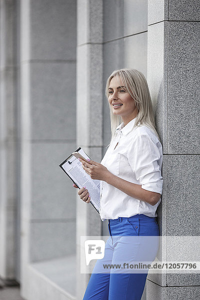 Smiling businesswoman with clipboard and cell phone leaning against a wall
