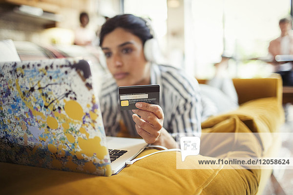Young woman with headphones and credit card online shopping at laptop on living room sofa