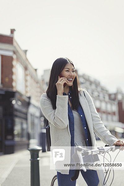 Lächelnde junge Frau  die mit dem Fahrrad pendelt  mit dem Handy auf einer sonnigen Straße in der Stadt telefoniert.