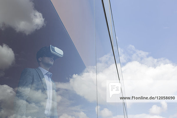Geschäftsmann mit Virtual-Reality-Simulator am modernen Bürofenster mit Blick auf blauen Himmel und Wolken