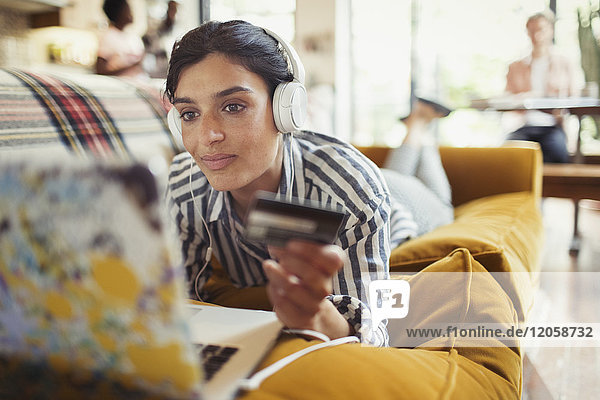 Junge Frau mit Kreditkarte und Kopfhörer online einkaufen am Laptop auf dem Wohnzimmersofa