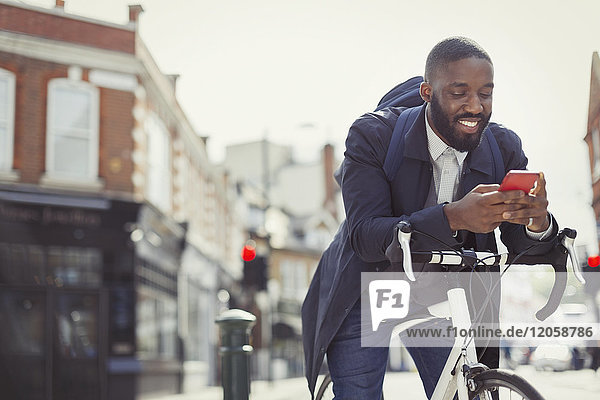 Junger Geschäftsmann  der mit dem Fahrrad pendelt  mit dem Handy auf der sonnigen Stadtstraße SMS schreibt