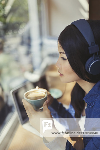 Pensive young woman drinking coffee  listening to music with headphones at cafe window