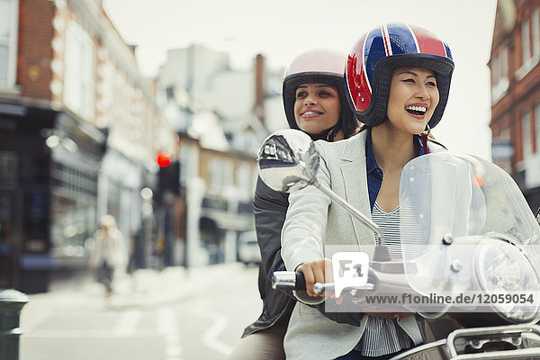 Lächelnde junge Frauen  die Helme tragen und Motorroller fahren.
