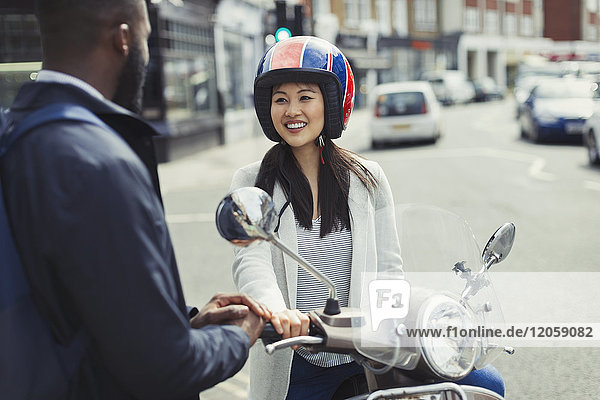 Lächelnde junge Frau auf dem Motorroller im Gespräch mit einem Freund auf einer sonnigen Stadtstraße.