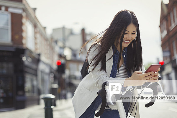 Smiling young woman commuting with bicycle  texting with cell phone on sunny urban street