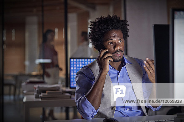 Businessman gesturing  talking on cell phone  working late at computer in dark office