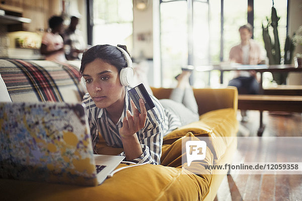 Young woman with headphones and credit card online shopping at laptop on living room sofa