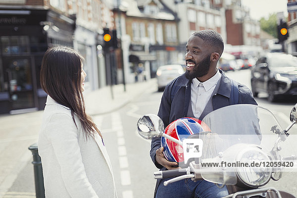 Lächelnder junger Geschäftsmann auf dem Motorroller im Gespräch mit einem Freund auf der Stadtstraße