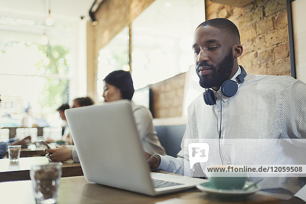 Junger Mann mit Kopfhörer über Laptop und Kaffeetrinken im Cafe