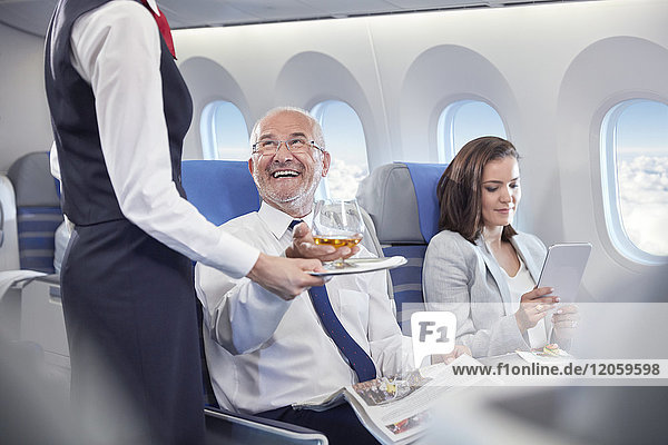 Flugbegleiter  der dem lächelnden Geschäftsmann Whiskey serviert  der in der ersten Klasse im Flugzeug sitzt.