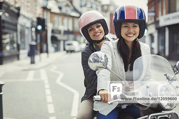 Lächelnde junge Frauen,  die Helme tragen,  Motorroller fahren in der Stadt.