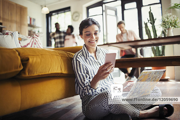 Lächelnde junge Frau mit Laptop und SMS mit Smartphone auf dem Wohnzimmerboden