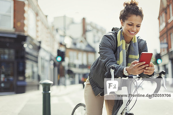 Lächelnde junge Frau  die mit dem Fahrrad pendelt  mit dem Handy auf der sonnigen Stadtstraße SMS schreibt