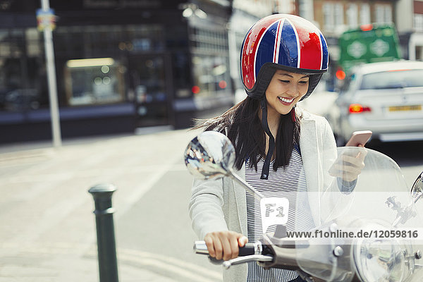 Lächelnde junge Frau beim SMSen mit Handy auf dem Motorroller  mit Helm auf der Straße.