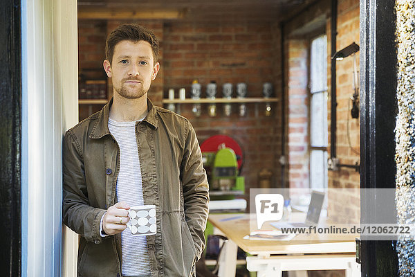Ein junger Mann steht an einer Werkstatttür und hält eine Kaffeetasse.