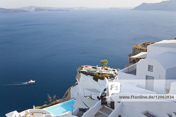 Hochwinkelaufnahme von traditionellen weiß getünchten Häusern und Swimmingpool auf einer griechischen Insel  in der Ferne ein Schiff im Mittelmeer.