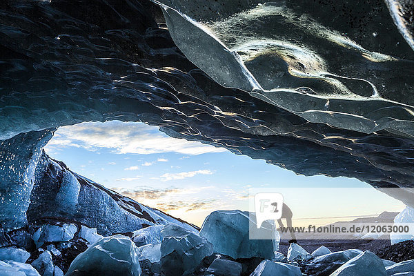 Side view of person climbing up on ice rock at the entrance to a glacial ice cave.