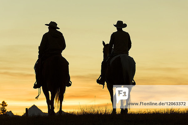 Zwei Cowboys reiten zu Pferd in einer Prärielandschaft bei Sonnenuntergang.