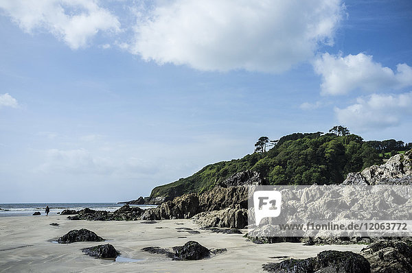 The headland and rocks on a sandy beach on the coastline under a blue sky with light cloud. A single person walking.