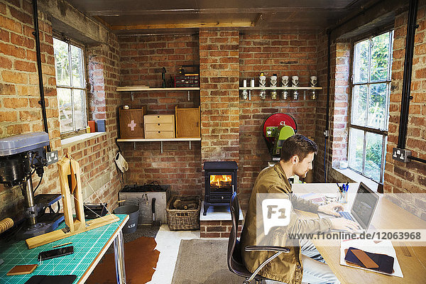 Ein Designer sitzt in seiner Lederwerkstatt an einem Schreibtisch mit einem Laptop. Ein Holzofen mit glühendem Feuer entzündet.