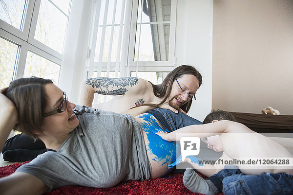 Eltern mit kleinem Sohn malen den Bauch der schwangeren Mutter mit blauer Farbe