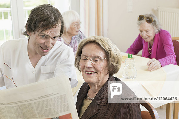 Caretaker reading newspaper with senior woman in rest home