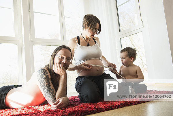 Portrait of parents sitting on carpet with little son measuring mother's pregnant belly