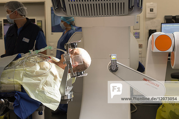 Reportage aus dem Operationssaal der stereotaktischen Neurochirurgie im Krankenhaus Pasteur 2  Nizza  Frankreich. Behandlung der Parkinson-Krankheit durch tiefe Hirnstimulation. Bei dieser Technik werden Elektroden in das Gehirn implantiert  die die abnorme elektrische Aktivität des Gehirns modulieren. Der Raum ist mit einem O-Arm-Scanner für die OP-Vorbereitung und dem NeuroMate  einem stereotaktischen Roboter  ausgestattet. Der Roboter ermöglicht es den Chirurgen  ihre Operationstechnik zu optimieren und zuverlässiger zu machen  und der O-Arm ermöglicht 3D-Bilder in Echtzeit  um die Präzision  Sicherheit und Effizienz der Operation zu verbessern. Der Beginn der Operation. Der Chirurg und sein Team bereiten mit dem O-Arm die Stellen vor.