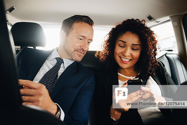 Smiling businessman colleagues sharing smart phone while sitting in taxi