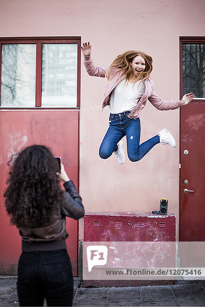 Rear view of teenage girl photographing female friend jumping against building