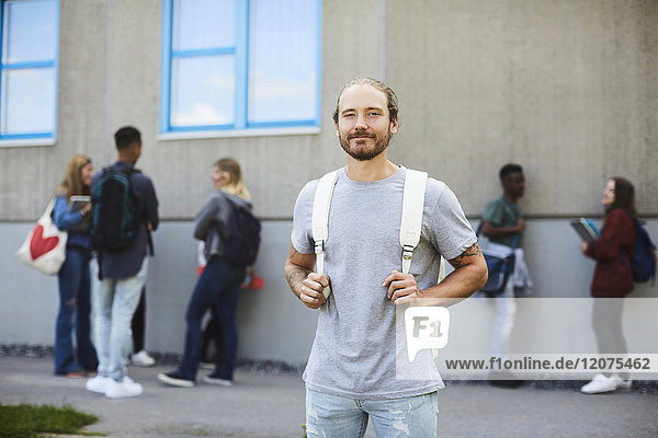 Porträt eines lächelnden Studenten mit Rucksack auf dem Universitätsgelände mit Freunden im Hintergrund